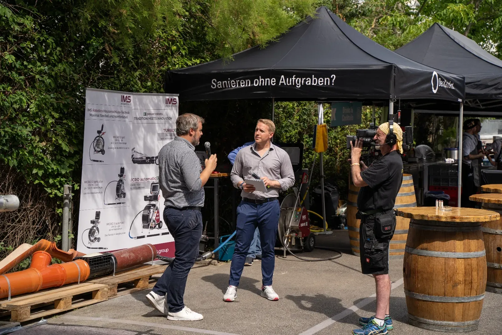 Interview vor der Kamera unter dem Pavillon „Sanieren ohne Aufgraben?“, daneben Rohre auf Paletten