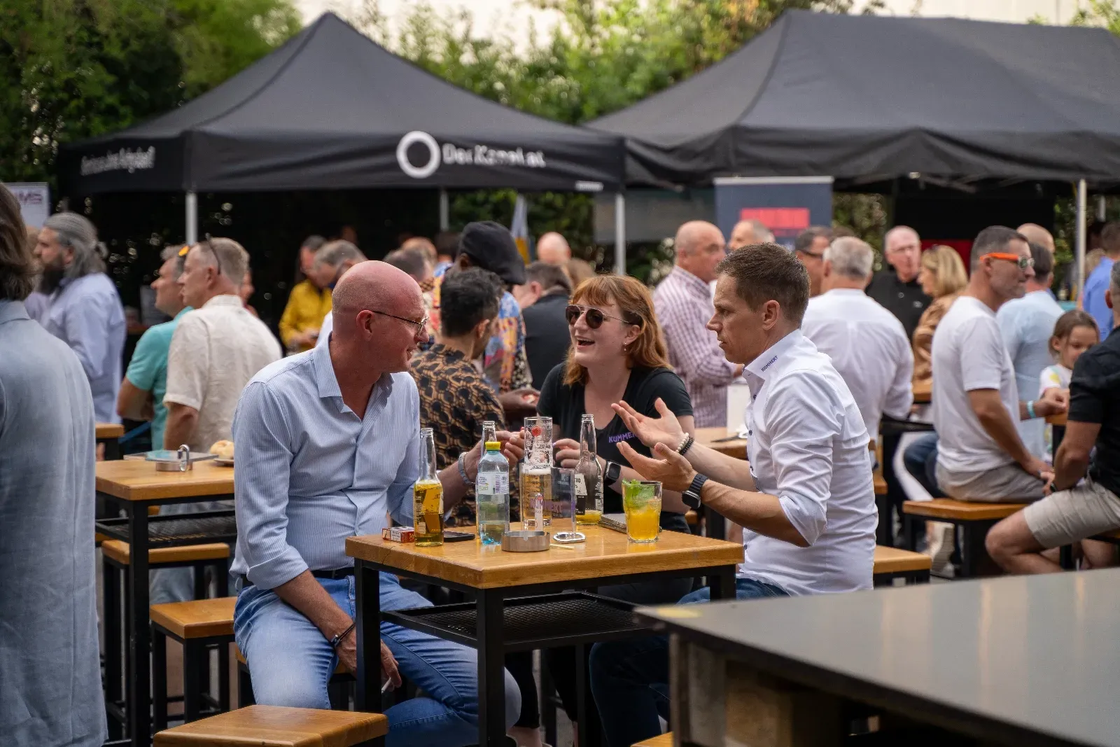 Drei Gäste im Gespräch am Stehtisch, im Hintergrund die volle Terrasse